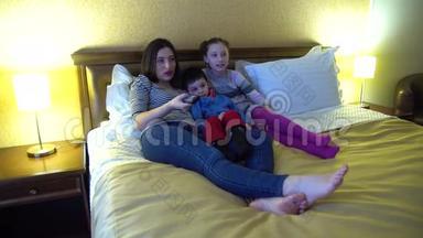一位年轻的母亲带着两个孩子，躺在旅馆的床上看电视。 使用电视遥控器。