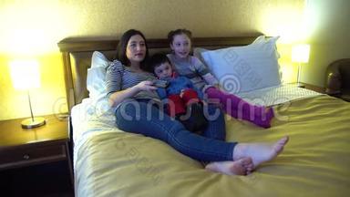 一位年轻的母亲带着两个孩子，躺在旅馆的床上看<strong>电视</strong>。 使用<strong>电视遥控器</strong>。