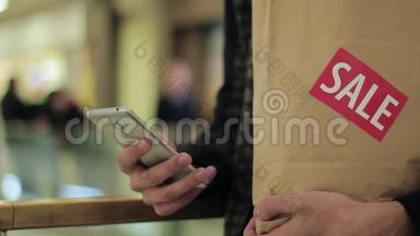 男士<strong>一只手</strong>拿着带有销售标签的棕色纸袋，在商场里使用智能<strong>手</strong>机