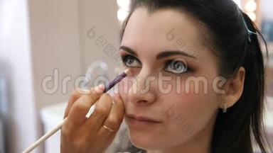 双手化妆师将化妆涂在一个年轻女人的脸上。 在专业沙龙里做眼妆的女孩。 晚上
