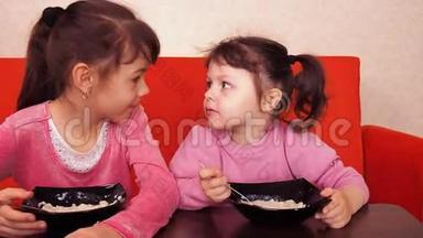 孩子们吃<strong>粥</strong>。 两个小女孩吃<strong>粥</strong>。 两个姐妹坐在橙色的沙发上吃晚饭。