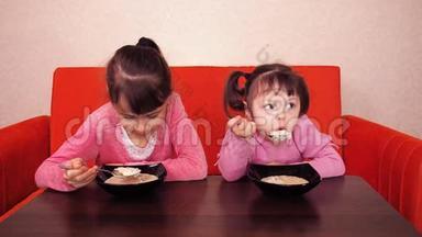 孩子们吃粥。 两个小女孩吃粥。 两个姐妹坐在橙色的沙发上吃晚饭。