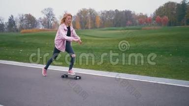 年轻漂亮的金发女郎穿着粉红色的风挡，玩滑板，滑着长板下山
