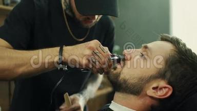 有才华的留胡子的理发师正在理发店里用黑色的<strong>剪发</strong>披风修剪他的客户的胡须。 他用的是