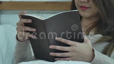生活方式快乐的年轻亚洲女人躺在床上看书，在家里穿着休闲服装。