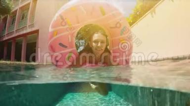 阳光明媚的夏日，戴着蓝色比基尼太阳镜的女人躺在充气的粉红色甜甜圈里漂浮在游泳池里。 看镜头