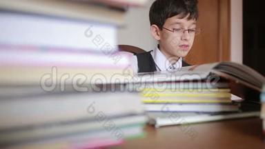 男孩坐在一张桌子旁，桌上放着一叠书。 孩子读书。 戴眼镜的男孩