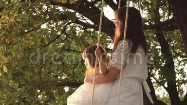 母亲在阳光下摇着女儿在树下摇摆。 特写镜头。 妈妈和宝宝坐在橡树树枝上的秋千上