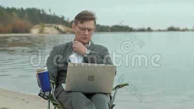 成熟的商人坐在河边放松。 穿西装用笔记本电脑的人。