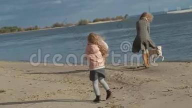 一个穿着外套的年轻女人和一个卷发女孩，妈妈和女儿，跑着，在海滩上和一只棕色的狗玩，狗
