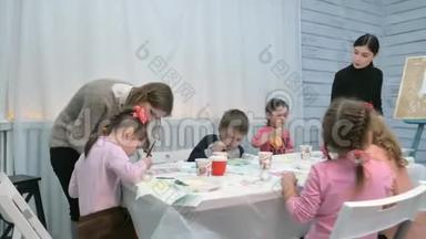 孩子们，男孩和女孩在教室里围着桌子坐在一起画画。 与他们在一起的是他们的年轻
