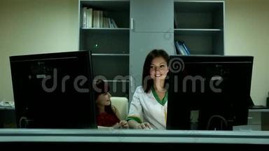 在商务办公室的两台电脑显示器后面，一个小可爱的女孩微笑着，一个年轻的黑发女医生