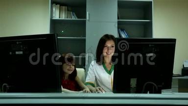 美丽的年轻黑发女医生在商务办公室对着两台电脑显示器后面可爱的小女孩微笑