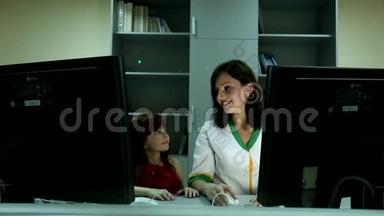 可爱的小女孩看着年轻漂亮的黑发女医生在商务办公室的两台电脑显示器后面微笑