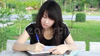 泰国学生青少年美丽的女孩写了一本书坐在公园里。