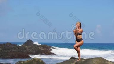 穿比基尼的年轻女孩一条腿站着做瑜伽，站在海滩上的岩石上，沙滩上有黑色的沙子