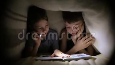 哥哥姐姐晚上在黑暗的房间里用手电筒在毯子下读一本书。 孩子们在玩。