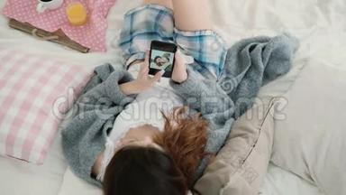 年轻漂亮的女人躺在床上拿着手机。 女孩在智能手机上看旅行的照片。