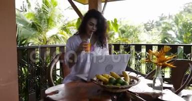 年轻女孩早餐时在夏季露台上使用笔记本电脑，美丽的女人在喝果汁时使用键盘