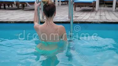 穿比基尼的迷人女人在蓝水背景下的梯子上从游泳池里爬出来，年轻的女人