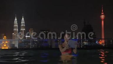 在摩天大楼屋顶上的游泳池里，女人的视线通过平板电脑拍摄夜间城市景观。 吉隆坡