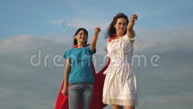 团队合作超级英雄。 两个穿着超级英雄红色<strong>斗篷</strong>的女孩站在蓝天上，风吹着<strong>斗篷</strong>。 妈妈