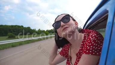 戴着太阳镜的快乐女孩斜靠在老式车窗外，享受旅行。 年轻女子望着窗外移动