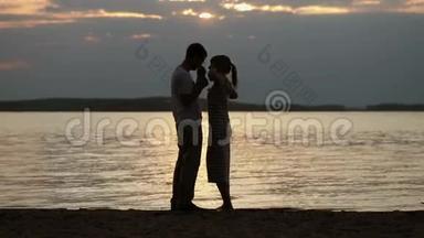 日落时分情人的剪影。 年轻漂亮的夫妇拥抱在湖岸上