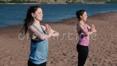 两个女人站在城市河边的海滩上伸展瑜伽。 美丽的城市景色。 令人讨厌的姿势。
