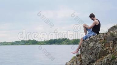 <strong>年轻人</strong>坐在一个乡村湖泊上方的岩石悬崖上。 乡村男孩。 孤独的<strong>年轻人</strong>