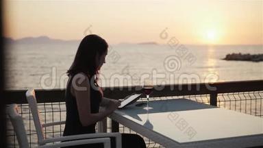日落时分坐在海边的咖啡馆里，年轻的布鲁内特用她的平板电脑端着一杯红酒