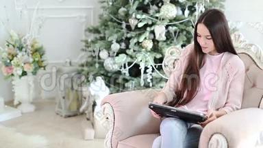 一个女孩坐在椅子上，在圣诞树下，用笔记本电脑工作。