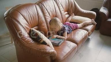 穿着粉色衣服的金发小女孩躺在沙发上，在平板电脑上玩游戏。 慢动作