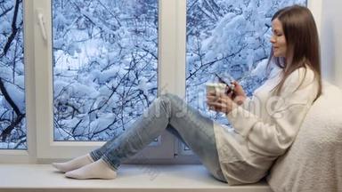 漂亮的女孩坐在窗台上，喝茶，用智能手机。 冬天在外面。