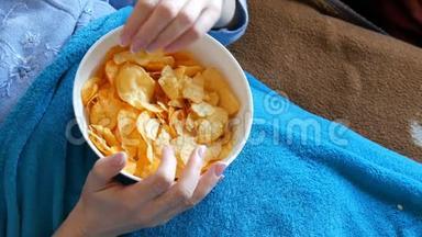 女人躺在蓝色毯子下的沙发上，吃着深盘子里的薯片。 不健康食品，快餐