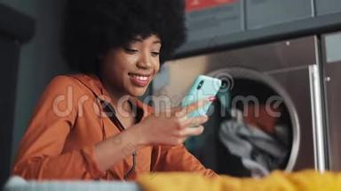 在洗衣店用智能手机微笑的年轻女人。 自助公共洗衣店。