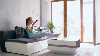 年轻漂亮的女人躺在沙发上看<strong>电视</strong>。 她无限期地打开<strong>遥控器</strong>，追踪镜头