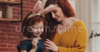 复活节快乐。 妈妈教女儿画鸡蛋。 为复活节做准备的幸福家庭。 他们戴着兔子耳朵
