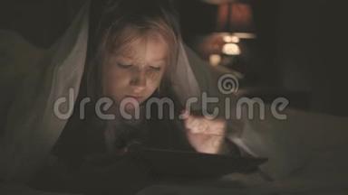 在黑暗的光线下，躺在床上的少女在社交网络上玩平板电脑。 特写小女孩看视频的画面