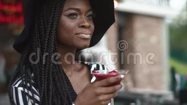 穿着黑色帽子和条纹时尚连衣裙的非裔美国女士的特写，酒吧里有清凉的鸡尾酒。 夏日炎炎