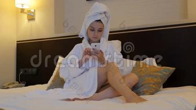 一个穿着白色外套的年轻漂亮的女人坐在酒店、卧室的床上，在电话里写短信。 这就是