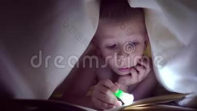 一个孩子晚上用手电筒在毯子下面读一本书。 有着淡发和蓝眼睛的男孩
