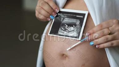 一位怀孕的年轻妇女在腹部拿着一张照片和一个怀孕测试。