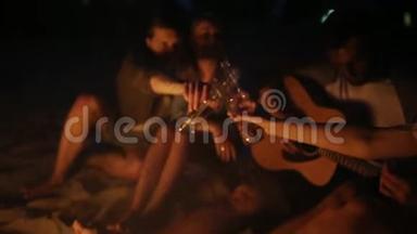 日落海滩派对，篝火晚会。 朋友们围坐在篝火旁，喝着啤酒，对着吉他唱歌。 青年