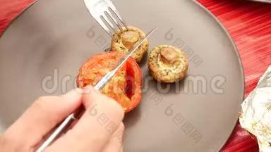 用锋利的不锈钢刀叉切烤番茄和蘑菇。 带有番茄和草菇的棕色圆形盘子