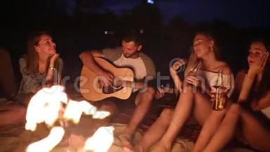 日落海滩派对，篝火晚会。 朋友们围坐在篝火旁，喝着啤酒，对着吉他唱歌。 男子和男子