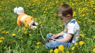 一个快乐的孩子和他的家人在蒲公英的春天的小路上。 一个带着手机的小男孩坐在绿色的草地上，上面有黄色的