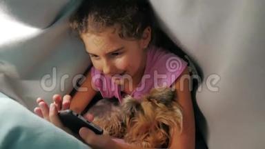 少女和狗在毯子下玩智能手机。 小女孩睡前躺在床上冲浪