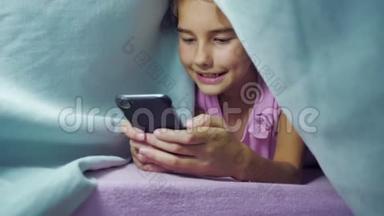 女孩少年和狗在毯子下的夜晚玩智能手机。 睡前的小女孩躺在床上冲浪