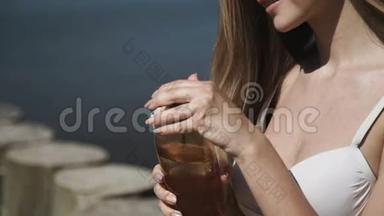 一个年轻的女人戴着夏帽，穿着白色<strong>比基尼泳衣</strong>，洗太阳浴，喝瓶子里的水。